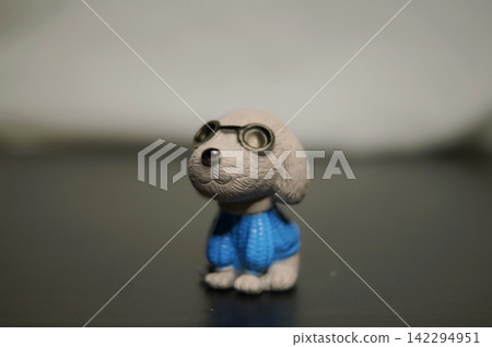 Small gray cartoon dog figurine wearing a blue outfit stands alone on a dark tabletop against a softly blurred background. The image represents individuality, confidence, companionship, and playful 142294951