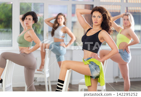 Experienced young female strip dancer dressed in casual activewear putting leg on chair and executing high heels dance moves with focused precision among group of people rehearsing in studio Experienced young female strip dancer dressed in casual activewear putting leg on chair and executing high heels dance moves with focused precision among group of people rehearsing in studio 142295252