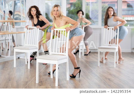 Positive smiling young woman and female group dancing strip plastic near chair in dance class, modern dance in stiletto heels 142295354