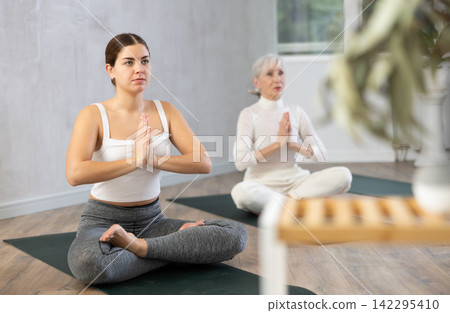 Young female yoga instructor showing others lotus pose in wellness center Young female yoga instructor showing others lotus pose in wellness center 142295410