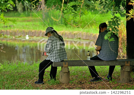 A person checking their smartphone in the shade near the pond next to the park. A person checking their smartphone in the shade near the pond next to the park. 142296251