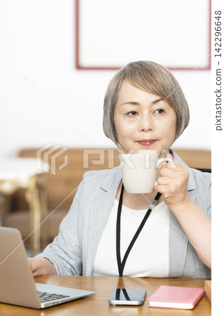 一位老年婦女在辦公室一邊工作一邊喝咖啡 一位老年婦女在辦公室一邊工作一邊喝咖啡 142296648