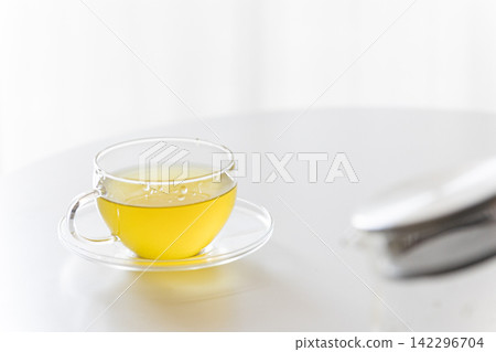 一張白色桌子上放著一個透明玻璃茶壺和一杯溫熱的綠茶/日本茶。 一張白色桌子上放著一個透明玻璃茶壺和一杯溫熱的綠茶/日本茶。 142296704