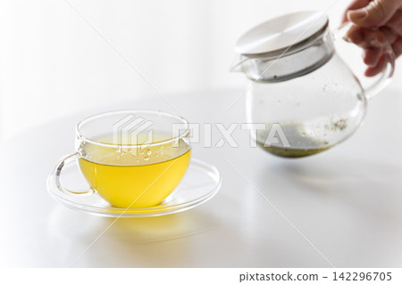 一張白色桌子上放著一個透明玻璃茶壺和一杯溫熱的綠茶/日本茶。 142296705