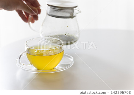 一張白色桌子上放著一個透明玻璃茶壺和一杯溫熱的綠茶/日本茶。 142296708