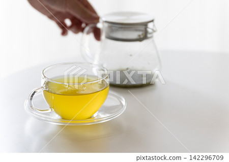 A clear glass teapot and a glass of warm green tea/Japanese tea are placed on a white table. A clear glass teapot and a glass of warm green tea/Japanese tea are placed on a white table. 142296709
