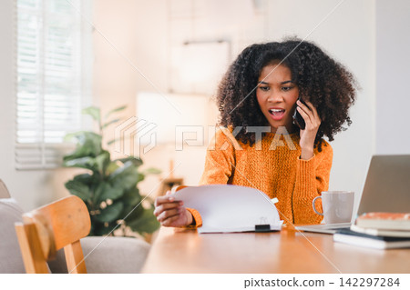 African American woman multitasking at desk, speaking on the phone while reviewing documents, with a laptop at home. African American woman multitasking at desk, speaking on the phone while reviewing documents, with a laptop at home. 142297284