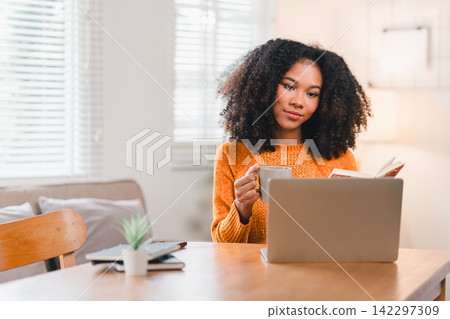 Smiling African American woman reading a notebook while working at home, sitting at a desk with a laptop and a coffee mug. Smiling African American woman reading a notebook while working at home, sitting at a desk with a laptop and a coffee mug. 142297309
