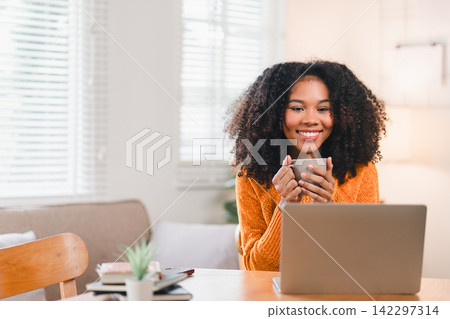 Happy African American woman holding a coffee mug while working on a laptop at home Happy African American woman holding a coffee mug while working on a laptop at home 142297314