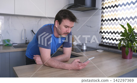 A Caucasian man stands at the kitchen table and looks at his mobile phone. He is dressed in a blue shirt and wears a neat beard and mustache A Caucasian man stands at the kitchen table and looks at his mobile phone. He is dressed in a blue shirt and wears a neat beard and mustache 142297369