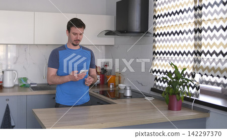 A lonely Caucasian guy stands in the kitchen with a phone in his hand and makes coffee in a turk. The kitchen is clean and well-furnished A lonely Caucasian guy stands in the kitchen with a phone in his hand and makes coffee in a turk. The kitchen is clean and well-furnished 142297370
