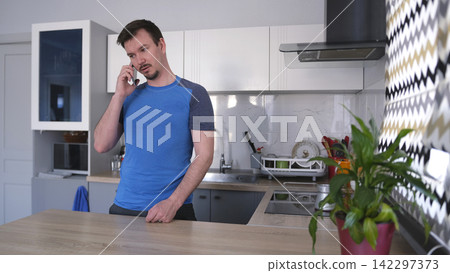 An attractive young man is talking on a mobile phone in a stylish kitchen. He makes coffee in a turk for breakfast. The kitchen is clean An attractive young man is talking on a mobile phone in a stylish kitchen. He makes coffee in a turk for breakfast. The kitchen is clean 142297373