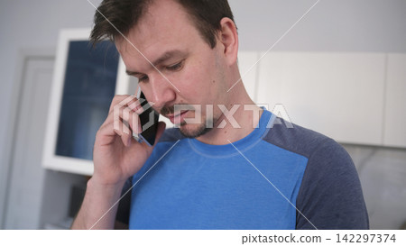 Close-up of a Caucasian man talking on a cell phone in the kitchen. He's a little upset and worried about the conversation. A call for work Close-up of a Caucasian man talking on a cell phone in the kitchen. He's a little upset and worried about the conversation. A call for work 142297374