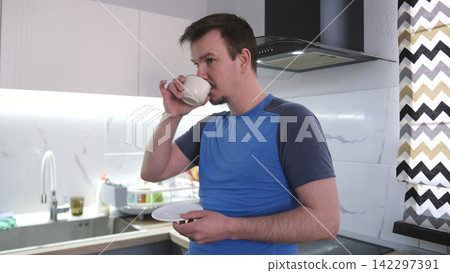 A cute young guy drinks fragrant coffee from a cup, holding a saucer in his hands. He bends his little finger aristocratically 142297391