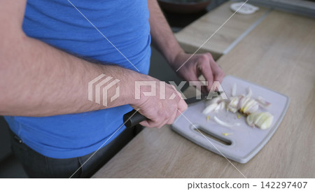 Close-up of an adult man chopping onions on a chopping board. The onion is cut into small pieces. The man is wearing a blue shirt Close-up of an adult man chopping onions on a chopping board. The onion is cut into small pieces. The man is wearing a blue shirt 142297407