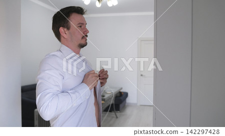 A Caucasian guy buttons up a white shirt while looking at himself in the mirror. A man gets dressed in front of a mirror for work A Caucasian guy buttons up a white shirt while looking at himself in the mirror. A man gets dressed in front of a mirror for work 142297428