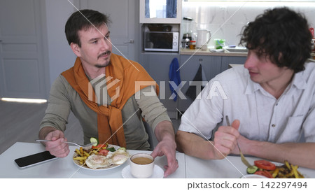 Two handsome young men are talking over a hearty lunch in a stylish kitchen. Gay men are discussing their wedding. Friends chat Two handsome young men are talking over a hearty lunch in a stylish kitchen. Gay men are discussing their wedding. Friends chat 142297494