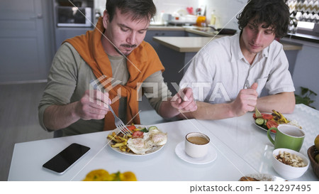 A cute curly-haired guy is looking at his friend's plate. Two men are having breakfast together at a table. A delicious lunch in an ordinary family A cute curly-haired guy is looking at his friend's plate. Two men are having breakfast together at a table. A delicious lunch in an ordinary family 142297495