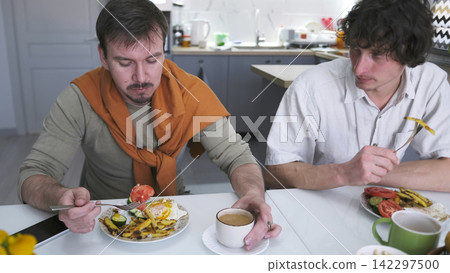 A family of young gay men quarrels over breakfast. Two men are eating at a table set with plates of food and cups of coffee 142297500