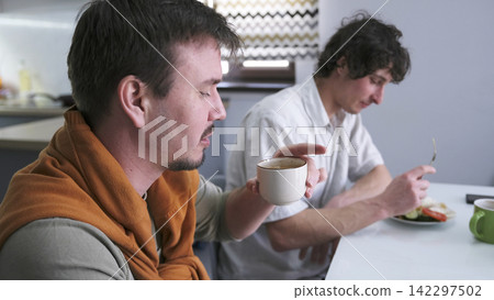 Lunch break. Two handsome young Caucasian men are sitting at a table in the kitchen. One drinks coffee and the other eats his lunch Lunch break. Two handsome young Caucasian men are sitting at a table in the kitchen. One drinks coffee and the other eats his lunch 142297502
