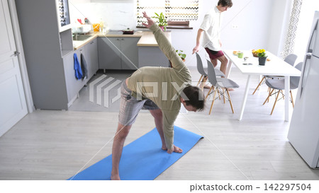 A guy exercises while his boyfriend has breakfast. A man takes a yoga pose on a blue rug in the kitchen. The ordinary life of gays A guy exercises while his boyfriend has breakfast. A man takes a yoga pose on a blue rug in the kitchen. The ordinary life of gays 142297504
