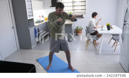 A young guy exercises while his roommate has breakfast. A man is working out in his living room with dumbbells A young guy exercises while his roommate has breakfast. A man is working out in his living room with dumbbells 142297506