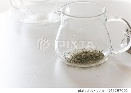 Green tea leaves in a transparent glass teapot placed on a white table. Green tea leaves in a transparent glass teapot placed on a white table. 142297601