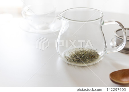Green tea leaves in a transparent glass teapot placed on a white table. Green tea leaves in a transparent glass teapot placed on a white table. 142297603