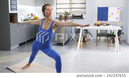 A pretty slender woman practices yoga lessons in the background of the kitchen. The student stands in a warrior pose. Health and sports A pretty slender woman practices yoga lessons in the background of the kitchen. The student stands in a warrior pose. Health and sports 142297635