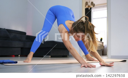 A woman takes a yoga pose in her living room. She's wearing a blue jumpsuit. A professional yogi. A young student practices yoga classes A woman takes a yoga pose in her living room. She's wearing a blue jumpsuit. A professional yogi. A young student practices yoga classes 142297638