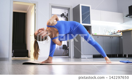 A sexy female yogi stands in a complex asana on a yoga mat. She practices oriental practices in her living room against the background of the kitchen A sexy female yogi stands in a complex asana on a yoga mat. She practices oriental practices in her living room against the background of the kitchen 142297650
