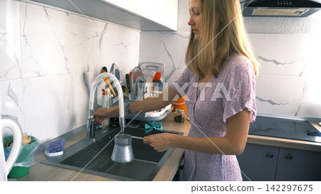 A pretty student draws water into a turk to make coffee on the stove. Household chores of a woman in the kitchen. Making coffee for breakfast A pretty student draws water into a turk to make coffee on the stove. Household chores of a woman in the kitchen. Making coffee for breakfast 142297675