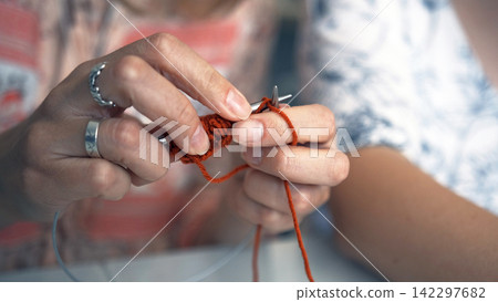 A young specialist knits a scarf made of red and orange yarn, close-up. Art and creativity. Knitting with knitting needles 142297682
