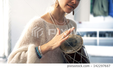 Close-up of a woman in a white knitted sweater playing a small drum. She is smiling and enjoying what is happening. Music and creativity Close-up of a woman in a white knitted sweater playing a small drum. She is smiling and enjoying what is happening. Music and creativity 142297687