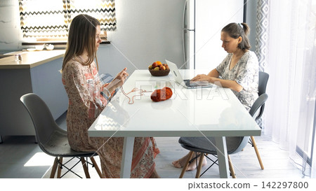 Two girlfriends are sitting at the kitchen table with a laptop and a bowl of fruit. One woman is typing on a laptop, and the other is crocheting Two girlfriends are sitting at the kitchen table with a laptop and a bowl of fruit. One woman is typing on a laptop, and the other is crocheting 142297800