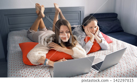 Two female students are lying on a bed with laptops in front of them. Two lesbians are smiling and enjoying their time together Two female students are lying on a bed with laptops in front of them. Two lesbians are smiling and enjoying their time together 142297815