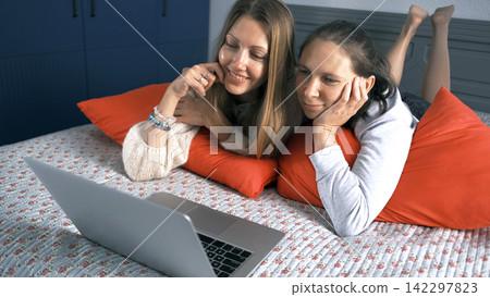 Two lovely happy women are lying on the bed, with a laptop in front of them. They look at the screen and smile Two lovely happy women are lying on the bed, with a laptop in front of them. They look at the screen and smile 142297823