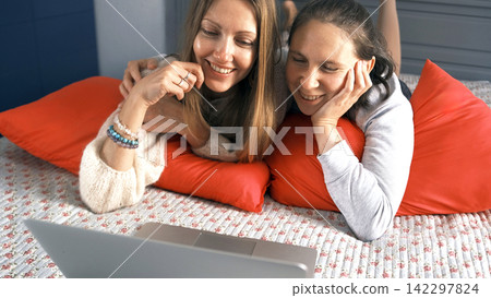 Two beauties are lying on the bed and laughing. One woman hugs another while watching a movie on a laptop Two beauties are lying on the bed and laughing. One woman hugs another while watching a movie on a laptop 142297824