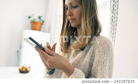 A woman is chatting enthusiastically on her phone. The blonde stands in the kitchen and looks at her mobile phone A woman is chatting enthusiastically on her phone. The blonde stands in the kitchen and looks at her mobile phone 142297825