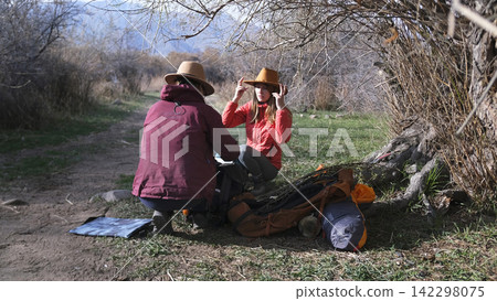 Trekking. Two people are sitting under a tree by a dirt road. The girlfriends look at the map to get to the right place. Hiking on a sunny day Trekking. Two people are sitting under a tree by a dirt road. The girlfriends look at the map to get to the right place. Hiking on a sunny day 142298075