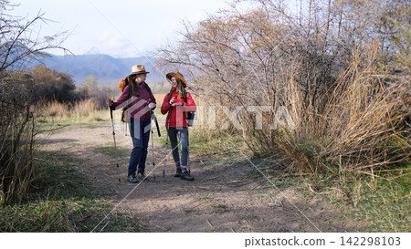 Two tourists are standing on a dirt road and chatting with each other. Two women with backpacks and trekking poles are relaxing after a long hike Two tourists are standing on a dirt road and chatting with each other. Two women with backpacks and trekking poles are relaxing after a long hike 142298103