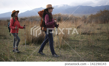 Two women are doing fitness in the field. Tourists with backpacks stroll through a grassy field against the backdrop of snow-capped mountains Two women are doing fitness in the field. Tourists with backpacks stroll through a grassy field against the backdrop of snow-capped mountains 142298143