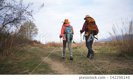 Two sisters hikers are walking towards the mountains. Weekend camping trip. Outdoor fitness. Hobbies of two students Two sisters hikers are walking towards the mountains. Weekend camping trip. Outdoor fitness. Hobbies of two students 142298148