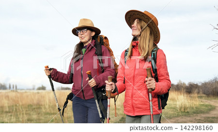 Two happy women in hats and backpacks are walking across the field. Travelers travel together. Outdoor fitness. Unity with nature 142298160