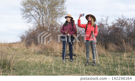 Two women are standing in a field, one of them is photographing beautiful landscapes on a mobile phone. Two travelers are walking in the wilderness Two women are standing in a field, one of them is photographing beautiful landscapes on a mobile phone. Two travelers are walking in the wilderness 142298164