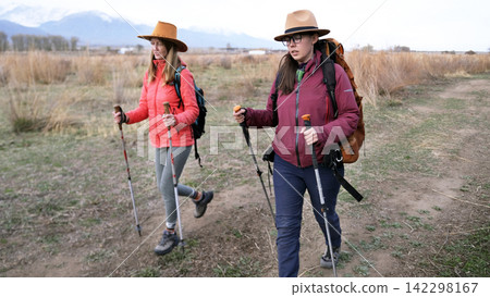 Two tired female students are walking along a dirt road with backpacks and hats. They have tracking sticks in their hands Two tired female students are walking along a dirt road with backpacks and hats. They have tracking sticks in their hands 142298167