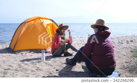 Two young students are sitting on the beach and drinking beer. There is a tent behind them. Women relax on the shore of the lake Two young students are sitting on the beach and drinking beer. There is a tent behind them. Women relax on the shore of the lake 142298290