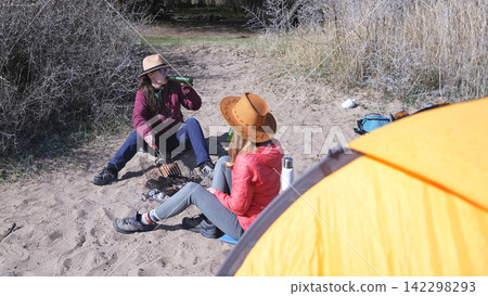 Two women are sitting on the beach next to a tent. Two alcoholics are having fun on the lake shore. Sisters roast sausages at a picnic Two women are sitting on the beach next to a tent. Two alcoholics are having fun on the lake shore. Sisters roast sausages at a picnic 142298293