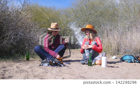 Two beauties are sitting on the beach by the campfire, one of them is frying sausages on a grill. Best friends are relaxing by the sea Two beauties are sitting on the beach by the campfire, one of them is frying sausages on a grill. Best friends are relaxing by the sea 142298296
