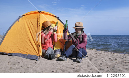 Two female students celebrate the end of the session. Women drink beer on the beach by the sea. Tourists are sitting by the tent and having fun Two female students celebrate the end of the session. Women drink beer on the beach by the sea. Tourists are sitting by the tent and having fun 142298301